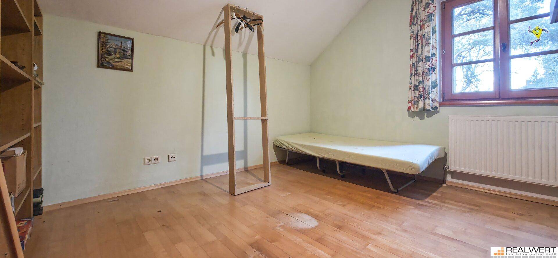 Einfaches Schlafzimmer mit Holzboden, einem Einzelbett und einem Fenster mit Blick ins Grüne.
