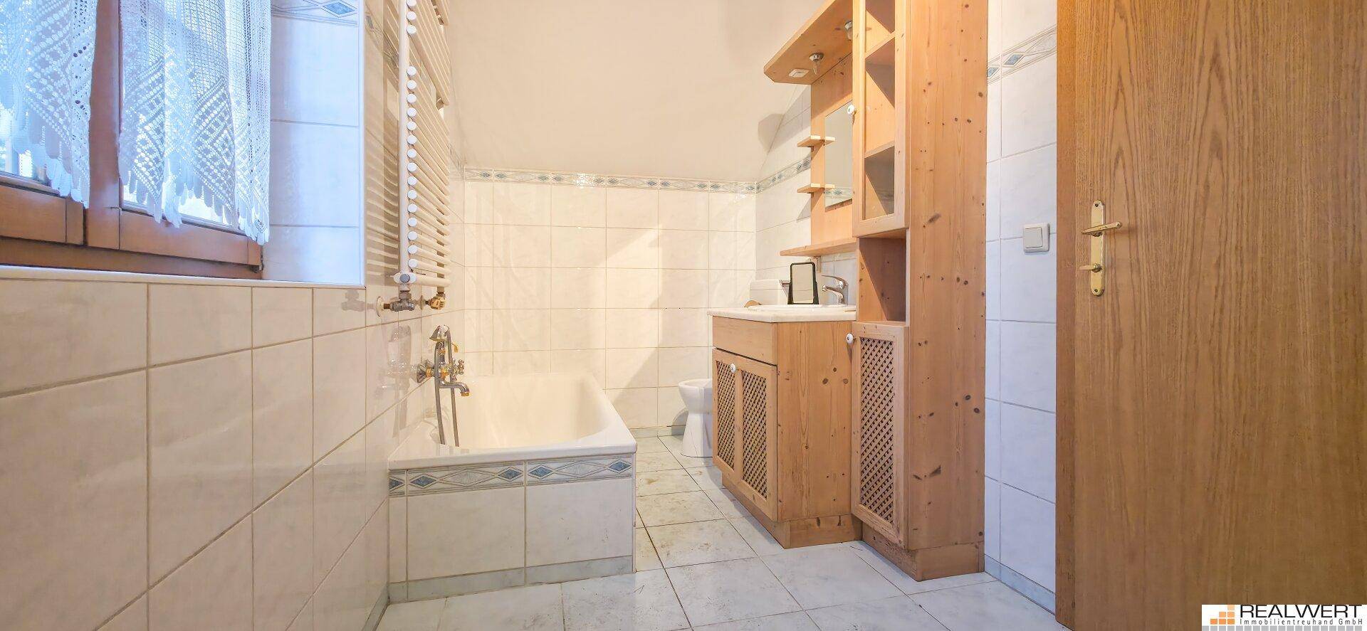 Helles Badezimmer mit Badewanne, weißen Fliesen und Holzmöbeln, ausgestattet mit einem Fenster.