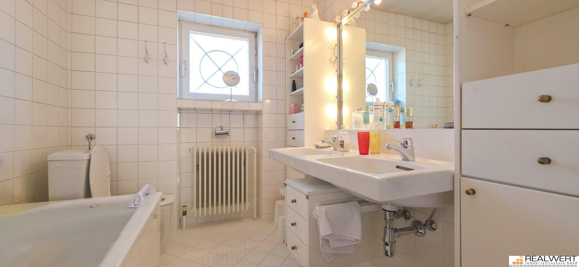 Helles Badezimmer mit weißen Fliesen, Badewanne, Doppelwaschbecken und Fenster.