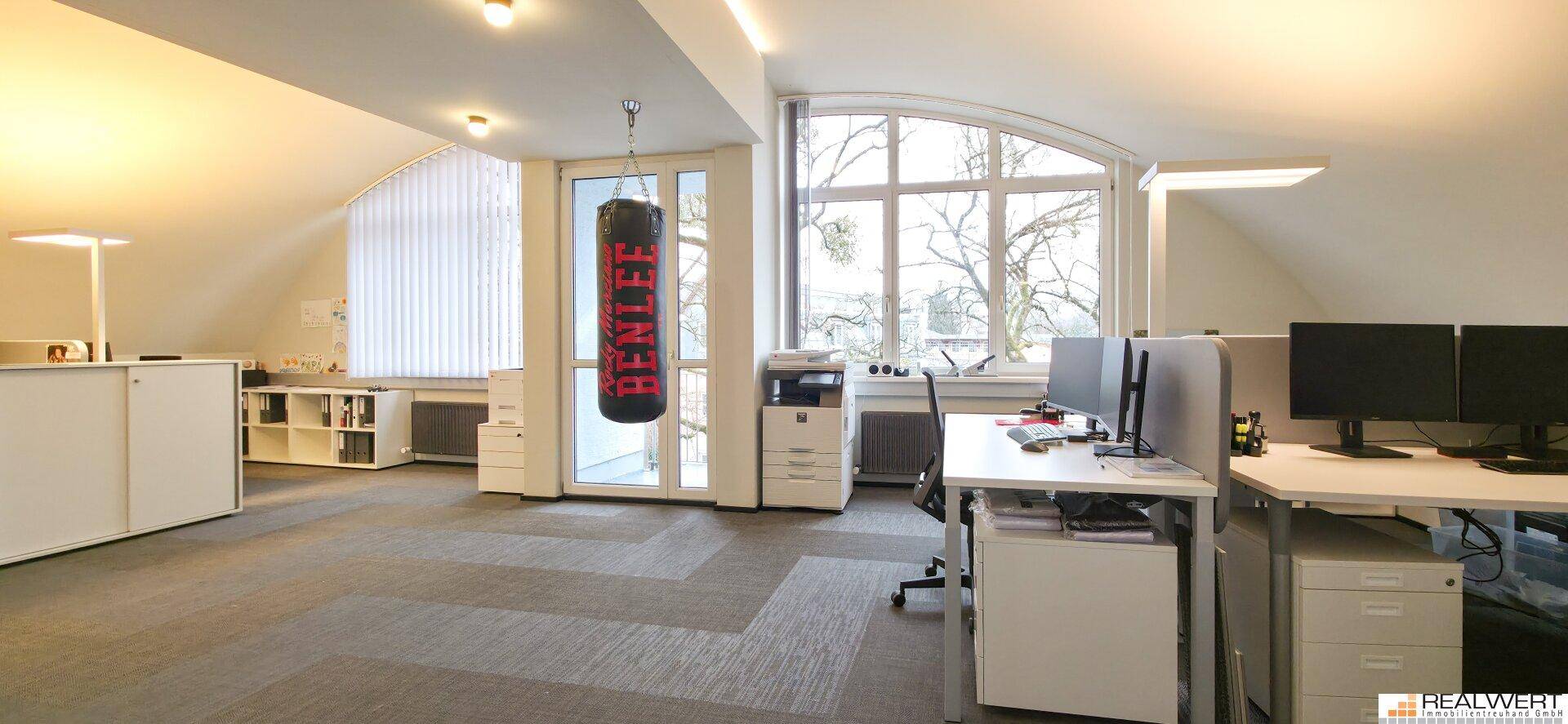Dachgeschossbüro mit großen Fenstern, schrägen Decken und mehreren Arbeitsplätzen.