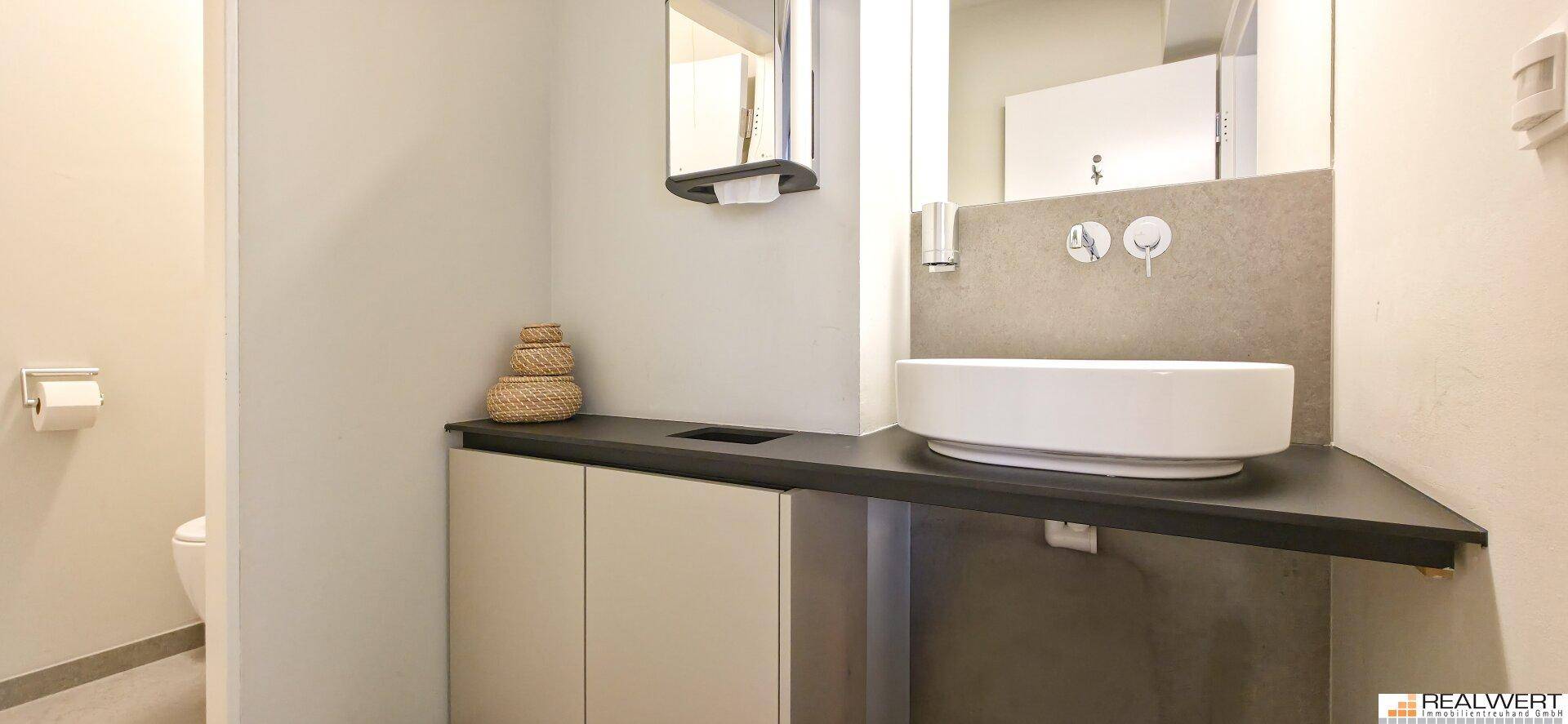 Modernes WC mit Aufsatzwaschbecken, dunkler Ablagefläche und hellem Fliesenboden.