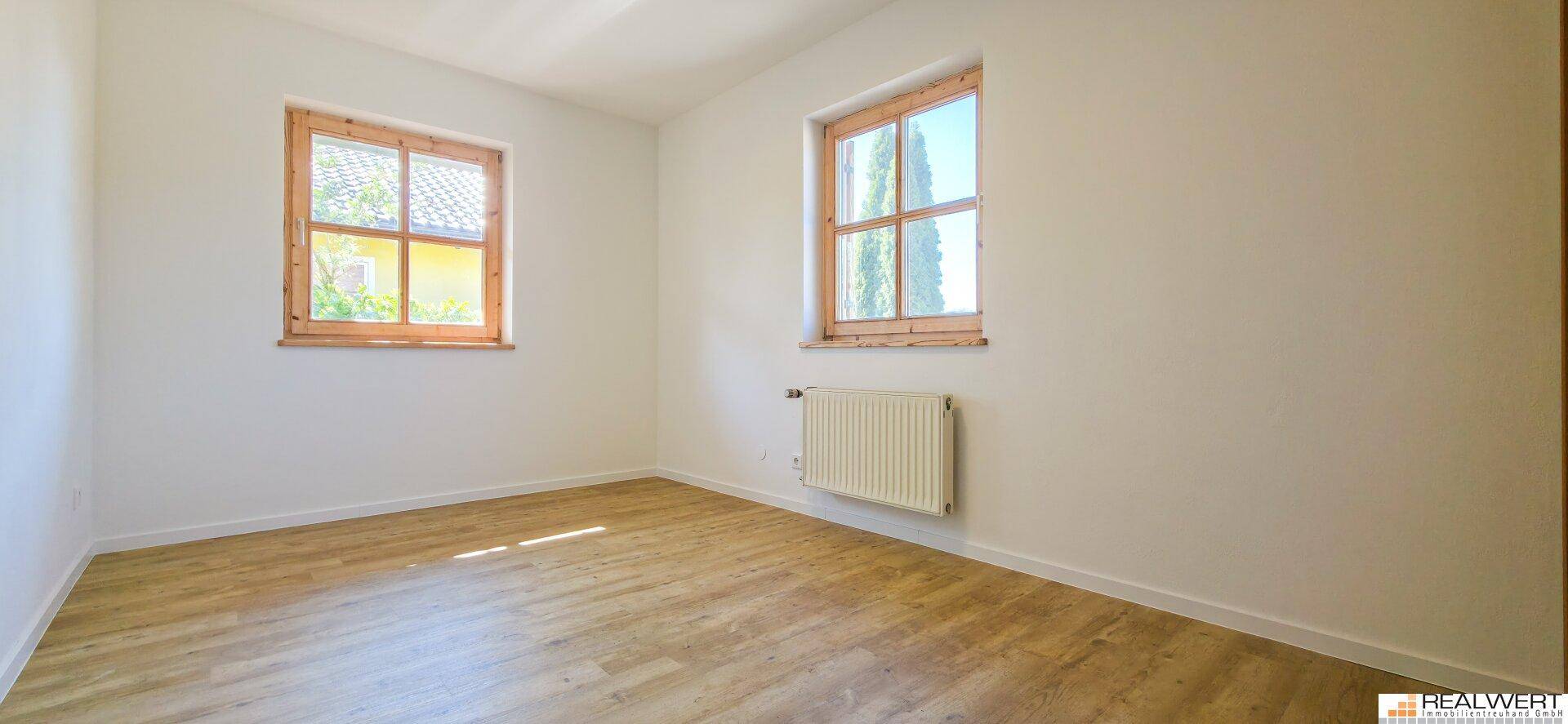 Helles, leeres Zimmer mit Holzboden und zwei Fenstern, ideal als Schlaf- oder Arbeitszimmer.