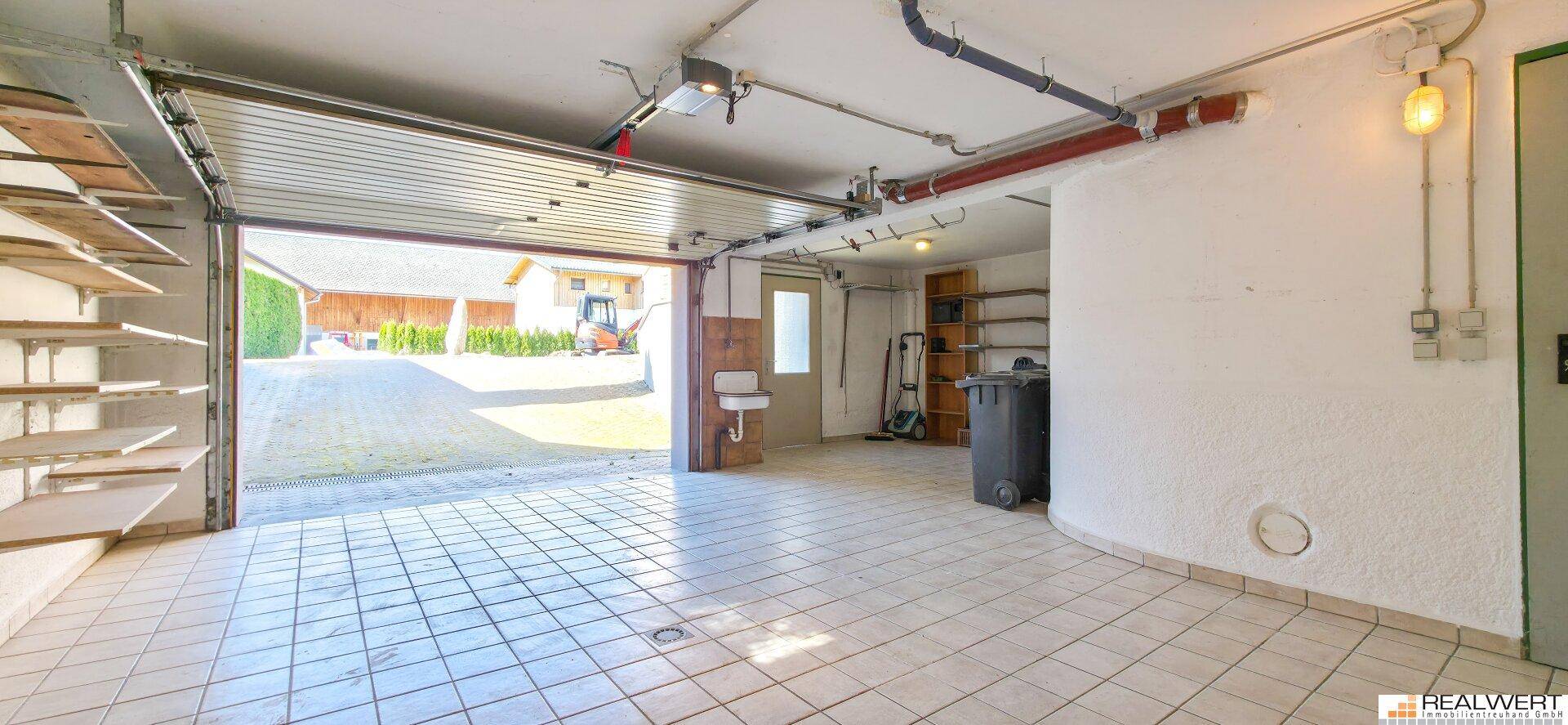 Geräumige Garage mit Fliesenboden und offenem Tor, das den Blick auf die Einfahrt freigibt.