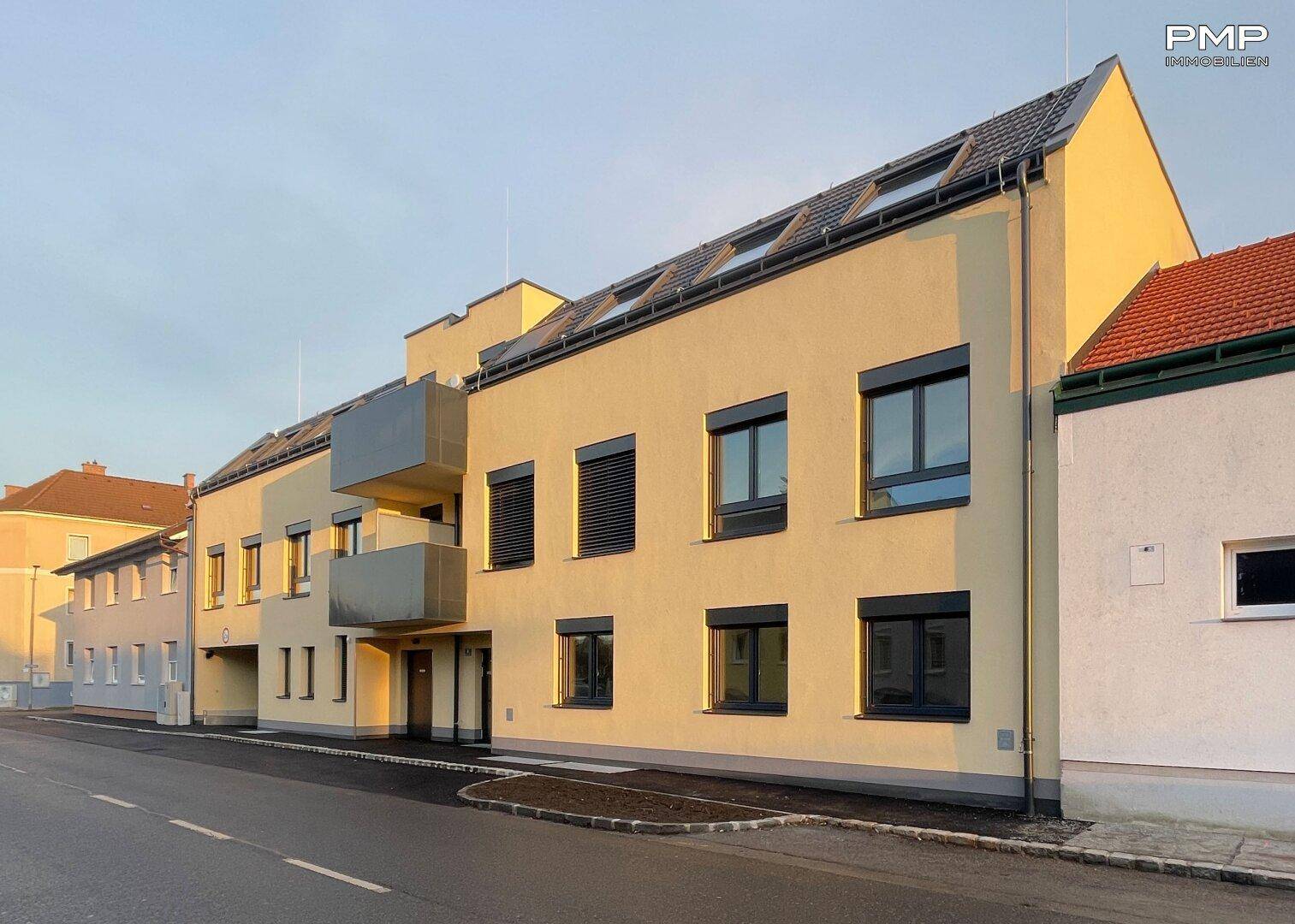 Modernes Mehrfamilienhaus mit heller Fassade, dunklen Fenstern und Dachgauben an einem sonnigen Tag.