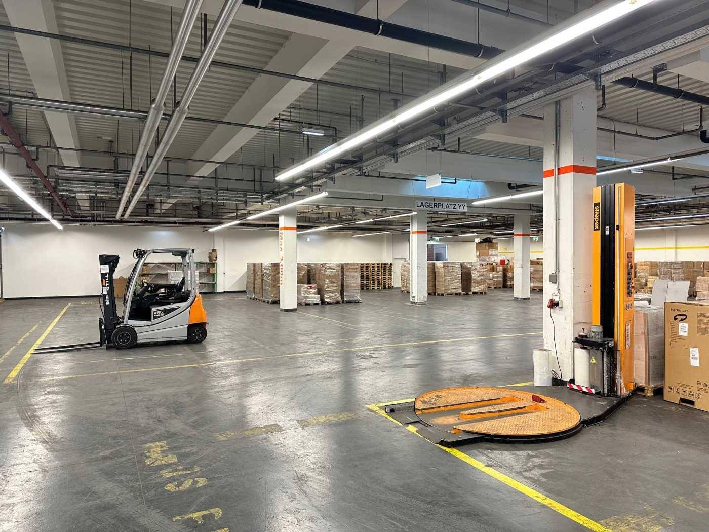 Weitläufige Lagerhalle mit Betonboden, einem Gabelstapler und einer Palettenwickelmaschine.