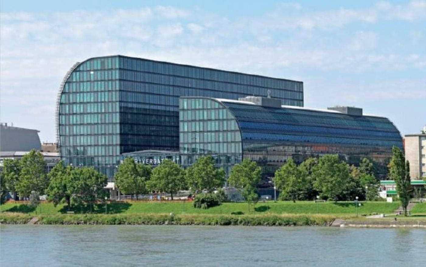 Moderne Bürogebäude mit geschwungenen Glasfassaden am Flussufer, umgeben von Bäumen.