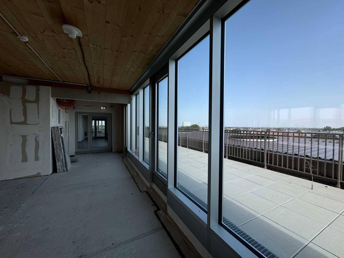 Langer Flur im Rohbau mit Holzbalkendecke und Zugang zu einer Außenterrasse mit Weitblick über die Stadt.