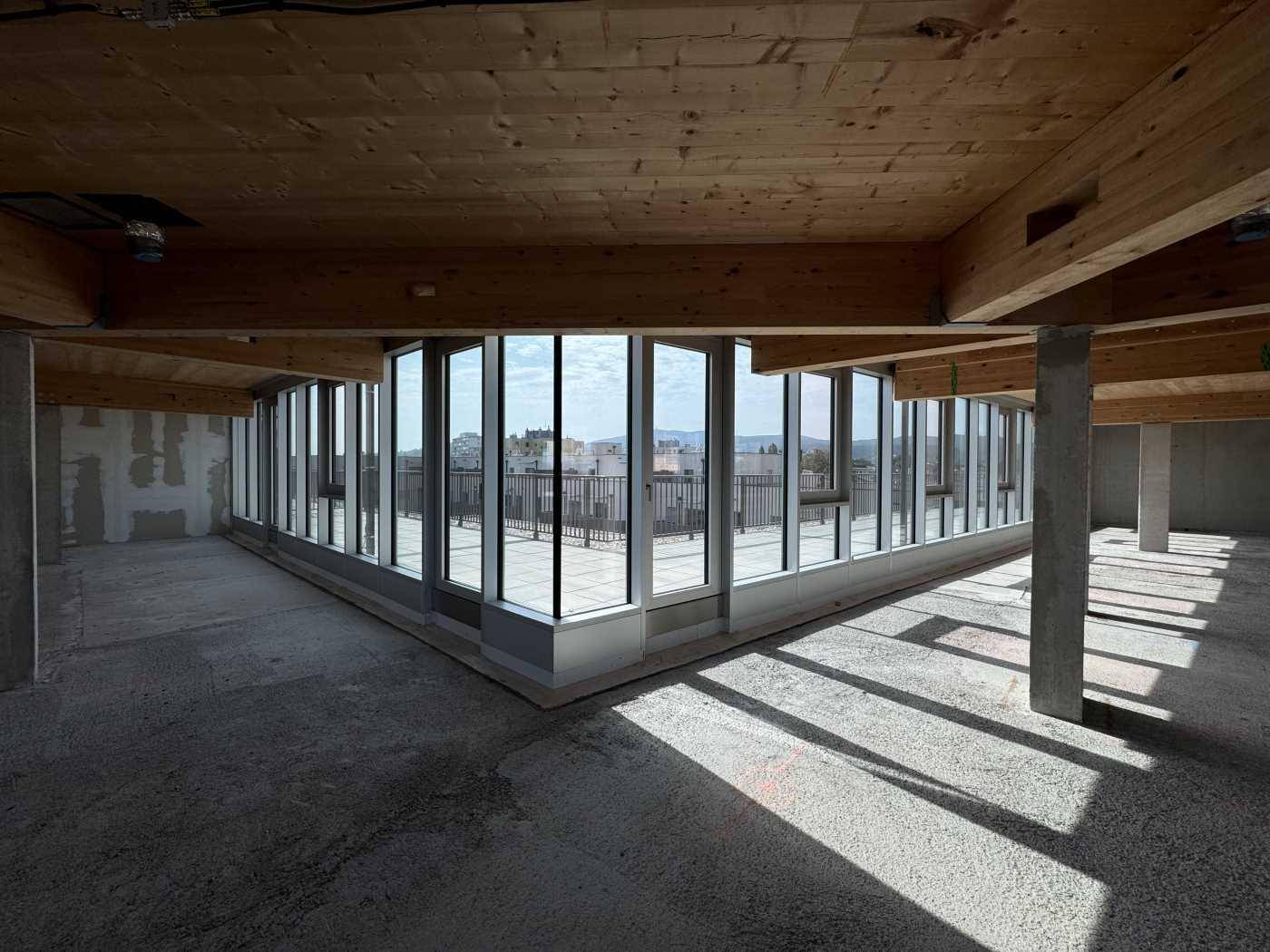 Heller Büroraum im Rohbau mit Holzbalkendecke und Eckfenstern, die viel Tageslicht hereinlassen und Weitblick bieten.