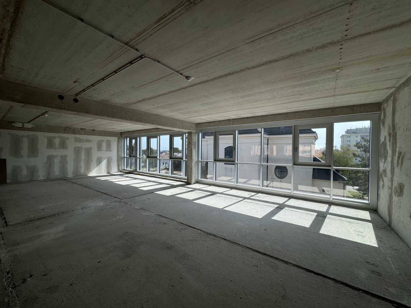Heller Büroraum im Rohbau mit Betonboden und großen Fenstern, die viel Sonnenlicht hereinlassen und Schatten werfen.