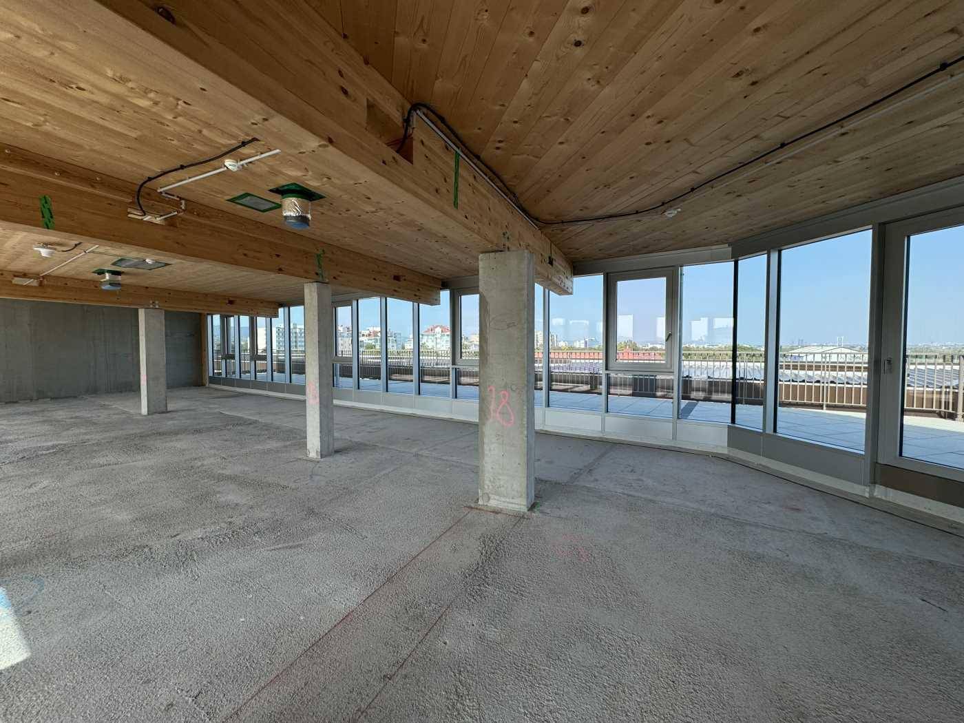 Großer Büroraum im Rohbau mit Holzbalkendecke, Betonstützen und Fensterfront mit Weitblick über die Dächer.