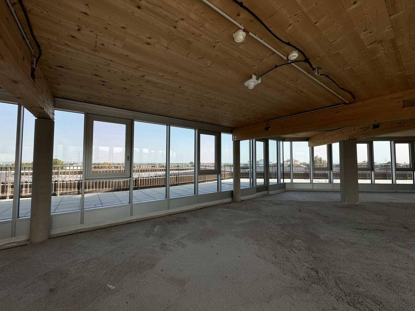 Geräumiger Büroraum im Rohbau mit Holzbalkendecke und Fensterfront mit Blick auf die Umgebung und den Horizont.