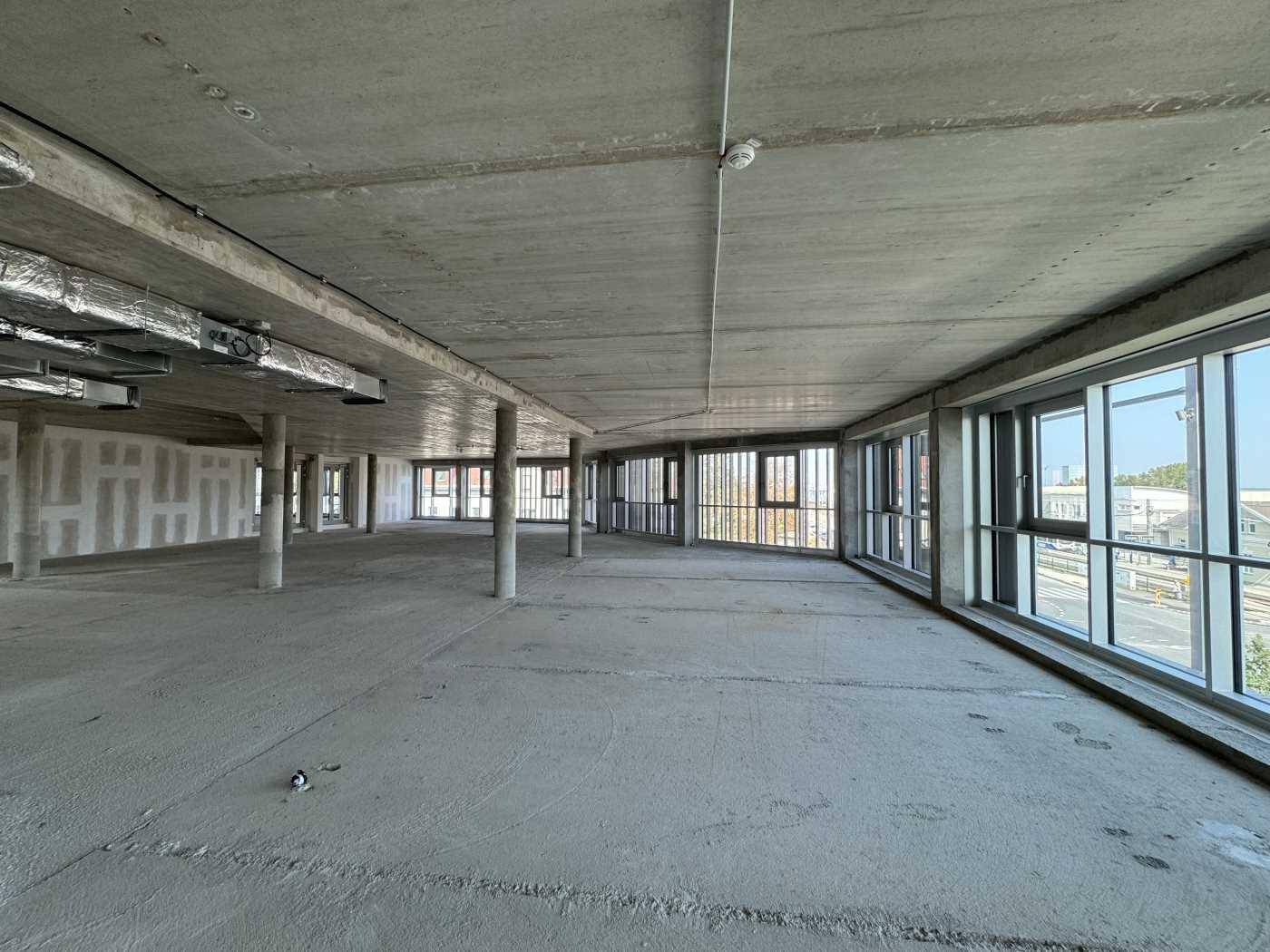 Weitläufiger Büroraum im Rohbau mit Betonboden, Säulen und großen Fenstern zur Straßenseite, bereit für den Ausbau.