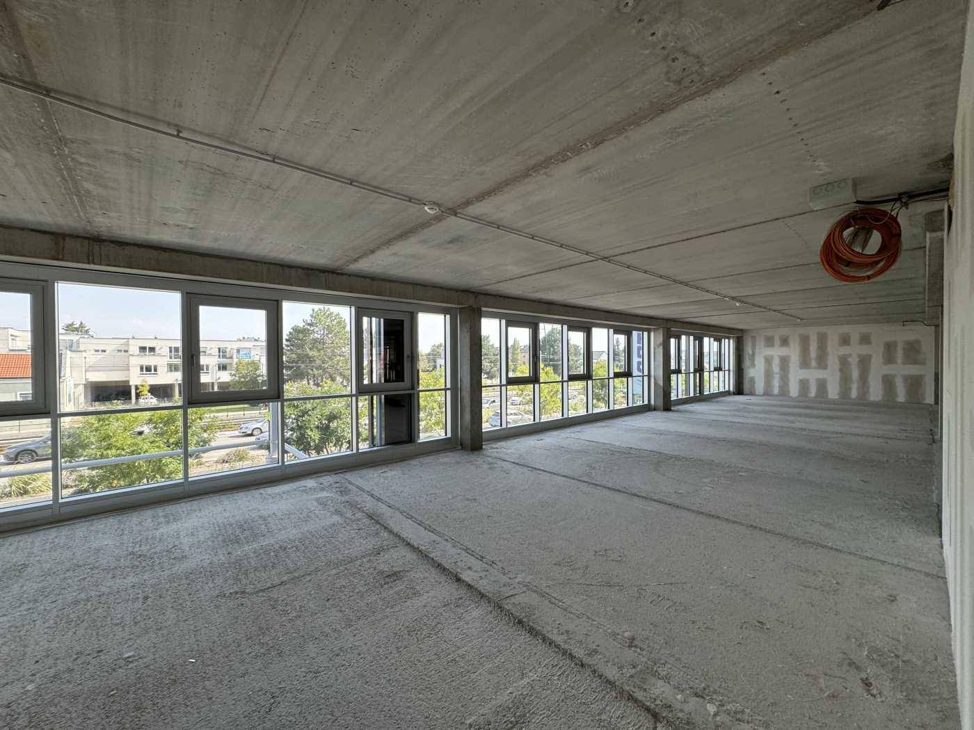 Großer, unfertiger Büroraum mit Betonboden und vielen Fenstern, die reichlich Tageslicht hereinlassen.