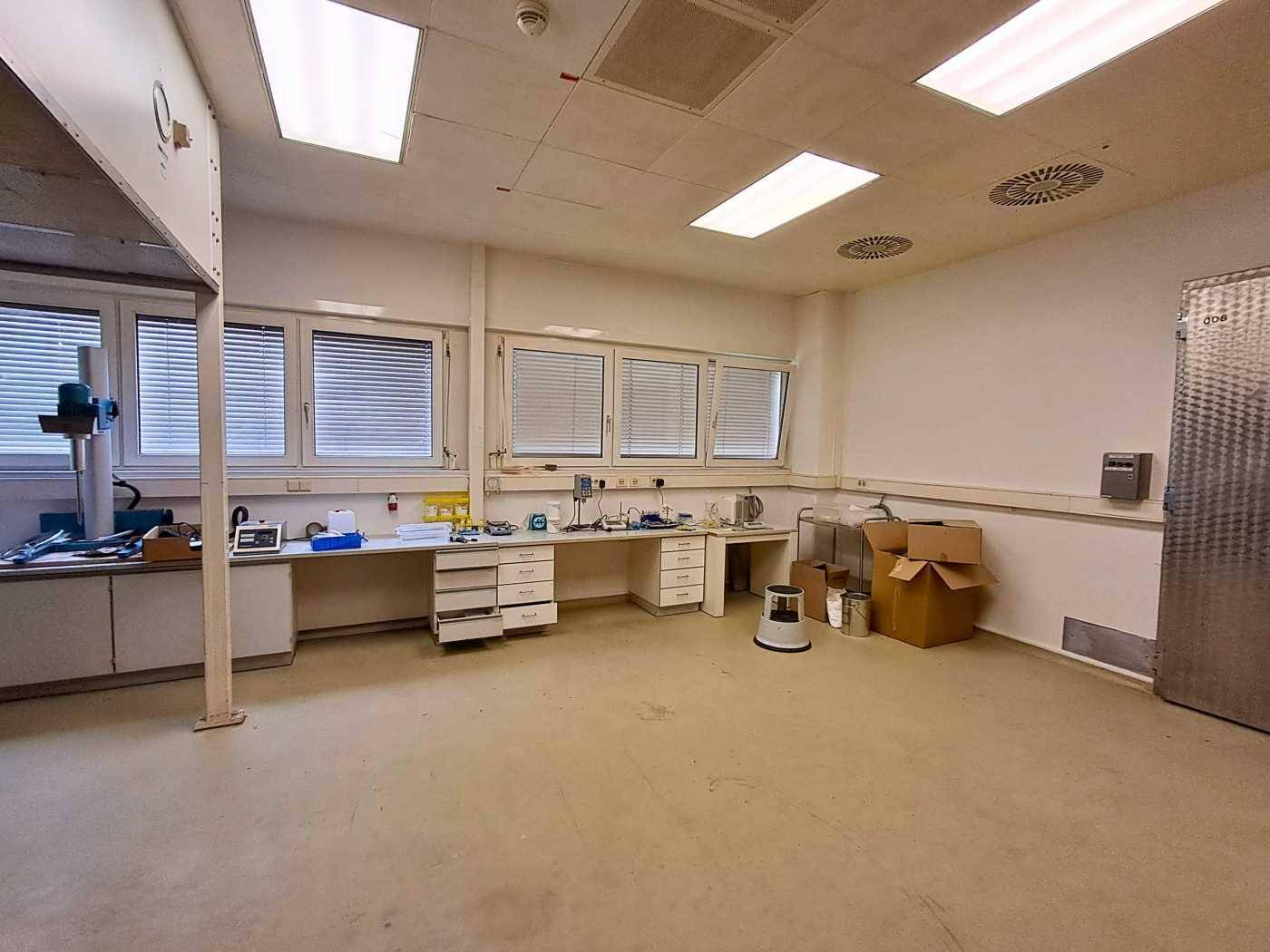 Heller Laborraum mit Arbeitsflächen, Geräten, großen Fenstern und Jalousien, geeignet für Forschung oder Werkstatt.