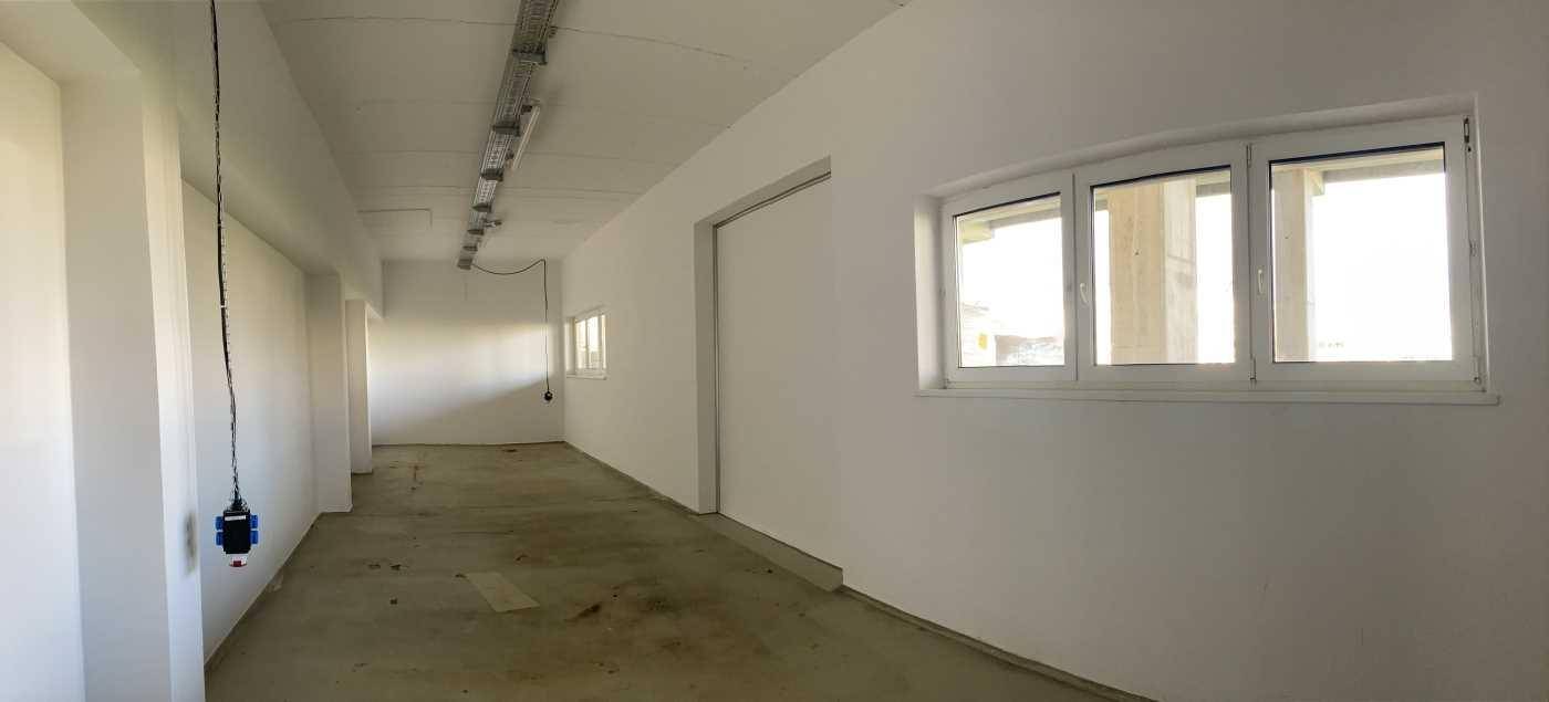 Langer, heller Raum mit weißen Wänden, großen Fenstern und sichtbaren Deckeninstallationen, geeignet als Flur oder Arbeitsbereich.