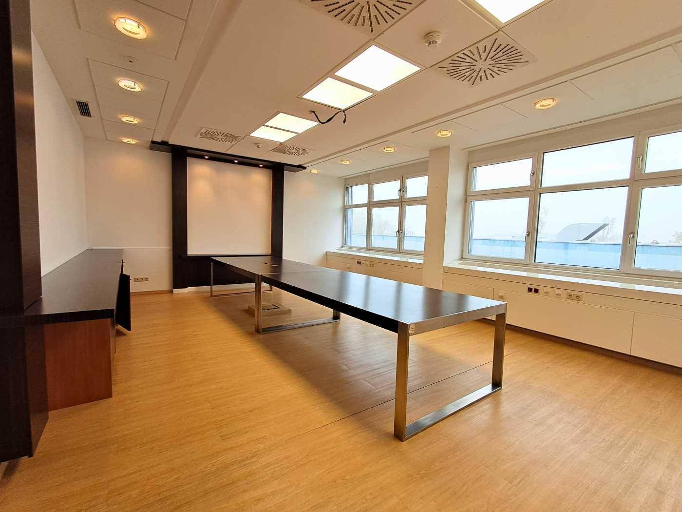 Moderner Konferenzraum mit großem Tisch, Projektionsfläche und hellen Fenstern.