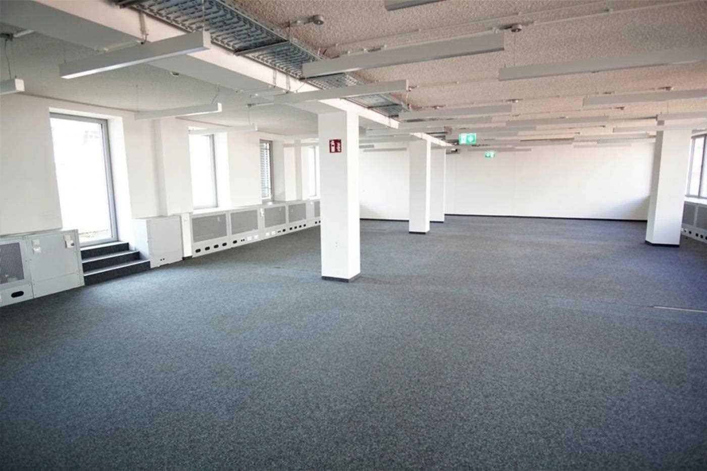 Helles, leeres Büro mit grauem Teppichboden, weißen Säulen und großen Fenstern mit Heizkörpern.