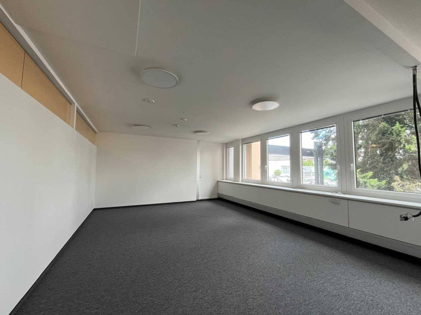 Heller, leerer Büroraum mit Teppichboden, großen Fenstern und einer abgehängten Decke, ideal für flexible Arbeitsplatzgestaltung.