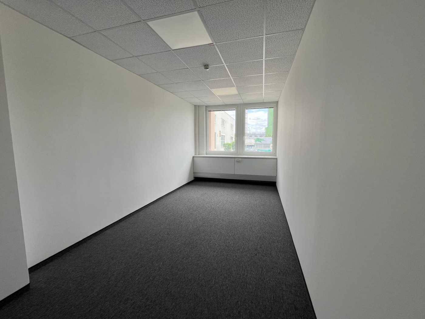 Langer, schmaler Büroraum mit Teppichboden und einem Fenster am Ende des Raumes.