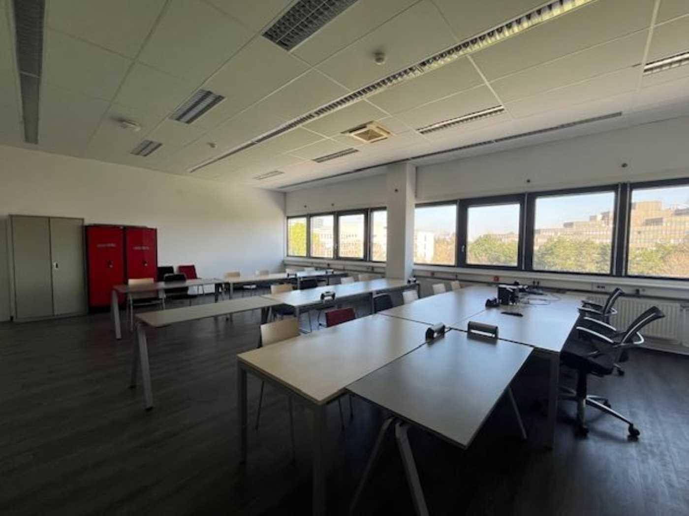Heller Schulungsraum mit mehreren Tischen, Stühlen und roten Spinden entlang der Wand.