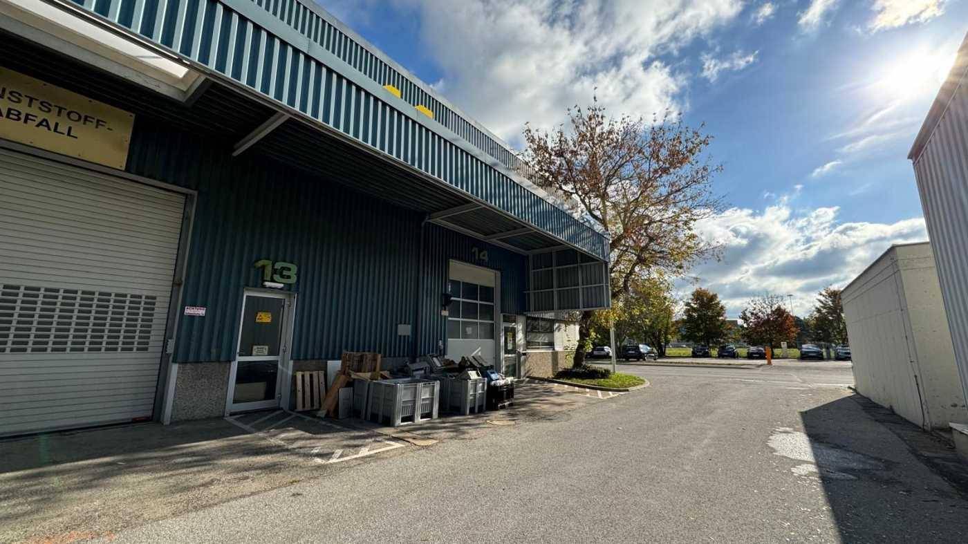 Industrielles Gebäude mit Rolltor und Überdachung, umgeben von einem Parkplatz und Bäumen.