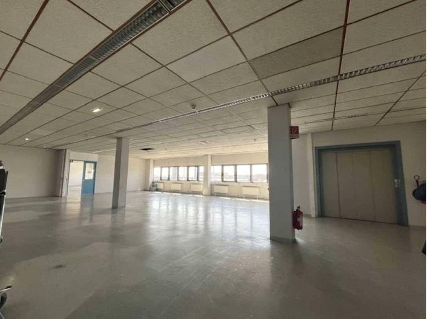 Großer, leerer Büroraum mit vielen Fenstern, Säulen und einem Aufzug im Hintergrund.