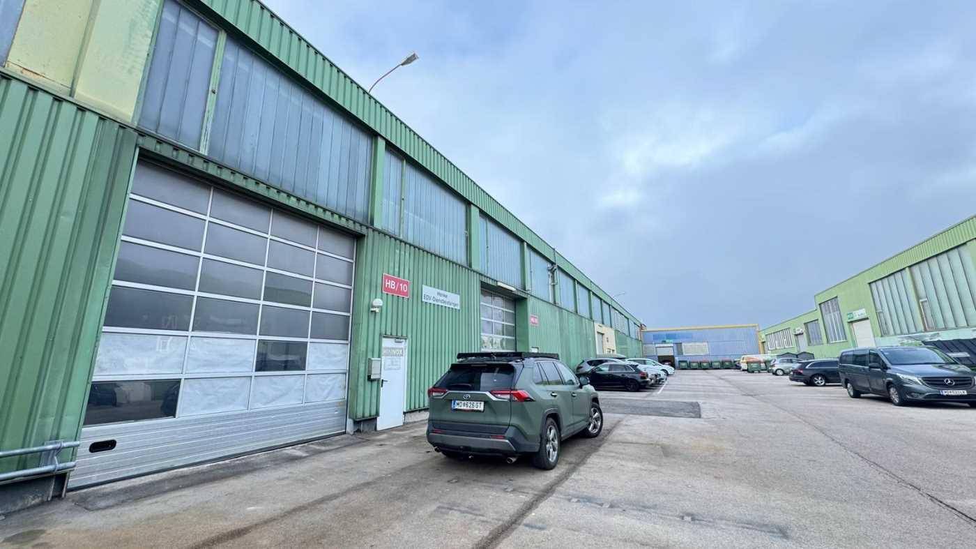 Grüne Industriefassade mit großen Rolltoren und Fenstern, davor ein großer Parkplatz mit mehreren Fahrzeugen.