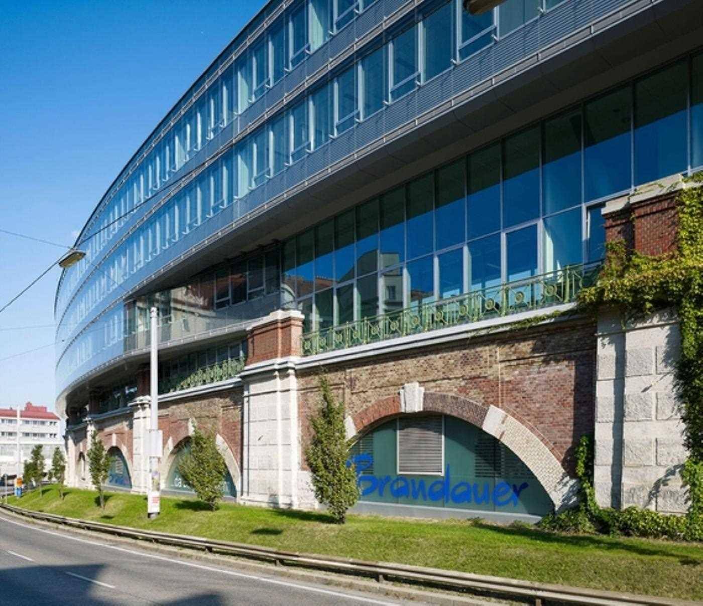 Moderne Glasfassade eines Bürogebäudes, die sich harmonisch mit historischen Backsteinbögen und der Straße verbindet.