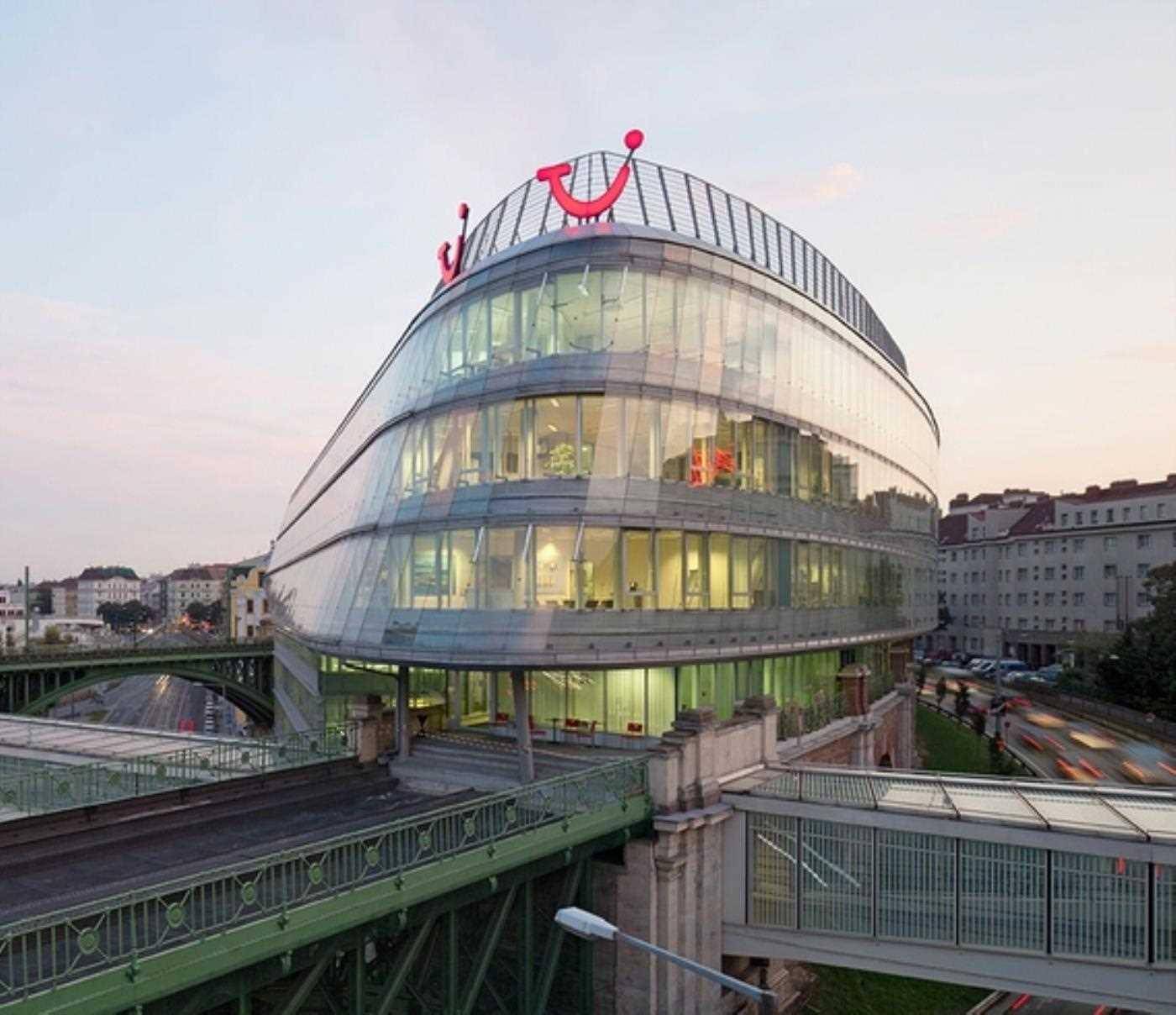 Geschwungenes Bürogebäude mit Glasfassade und TUI-Logo bei Sonnenuntergang, umgeben von städtischer Infrastruktur.