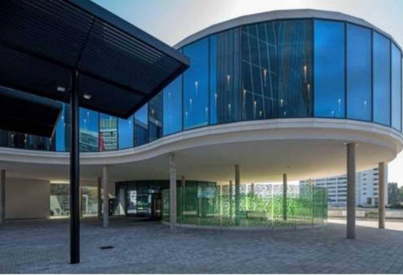 Modernes Gebäude mit geschwungener Glasfassade und überdachtem Eingangsbereich, umgeben von Grünflächen.