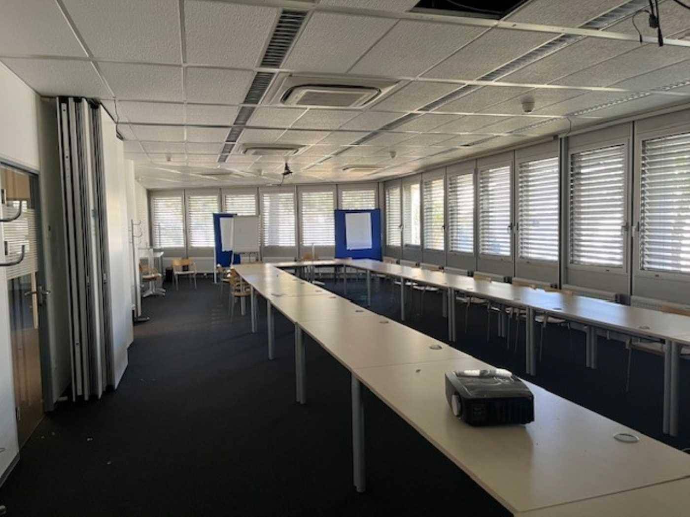 Großer Konferenzraum mit U-förmiger Tischordnung, vielen Fenstern und einem Projektor für Präsentationen.