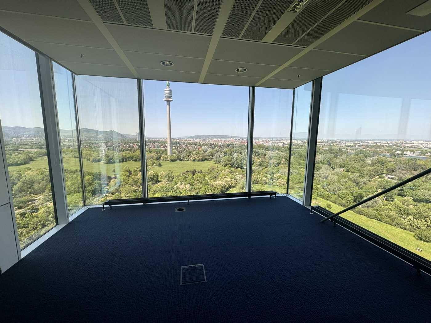 Eckbüro mit raumhohen Glaswänden und atemberaubendem Panoramablick auf grüne Landschaften und den Donauturm.