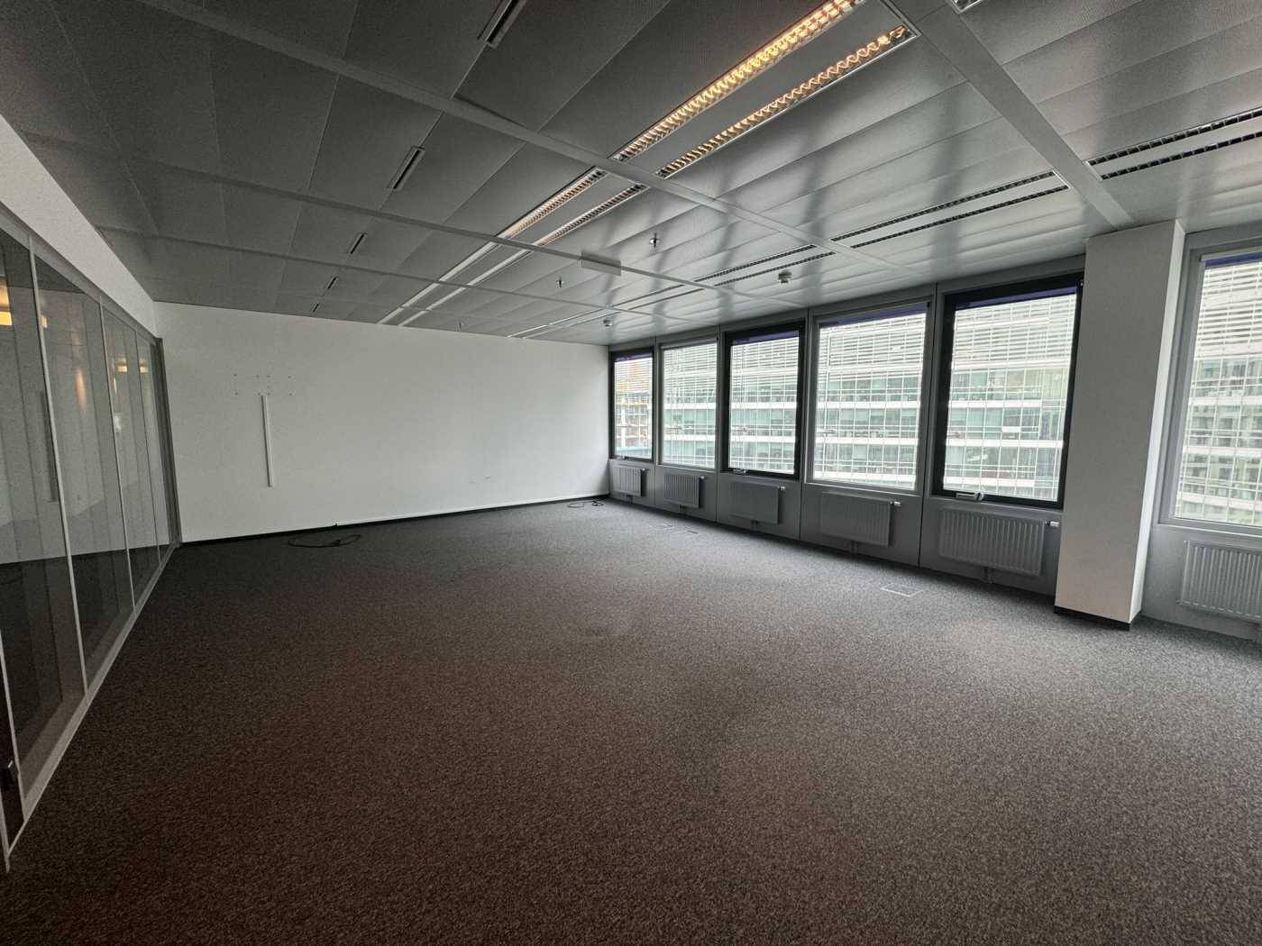 Geräumiges, leeres Büro mit Teppichboden, abgehängter Decke und großen Fenstern mit Stadtblick.