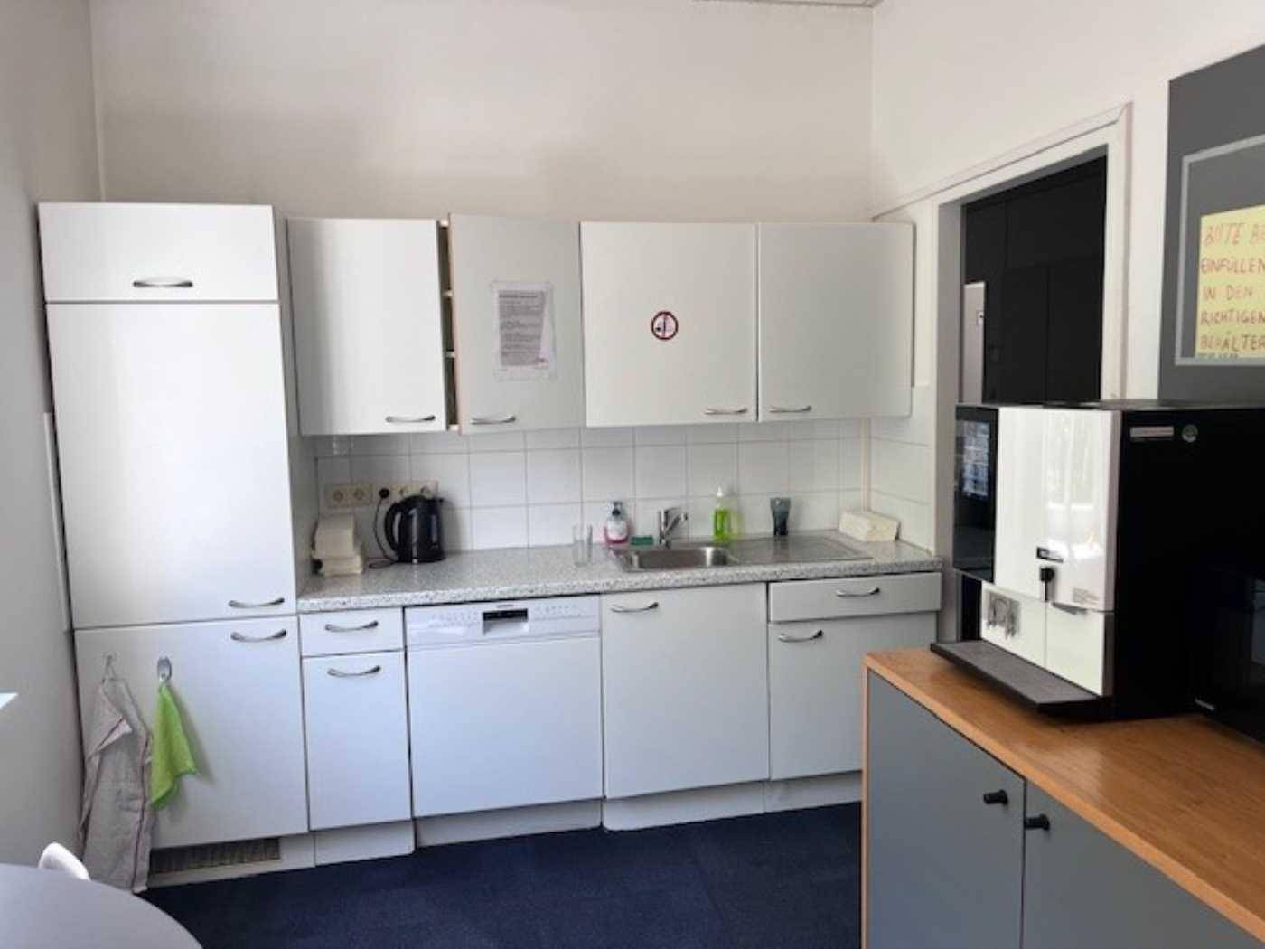 Funktionale Teeküche mit weißen Schränken, Spülmaschine und Kaffeemaschine für Mitarbeiter.