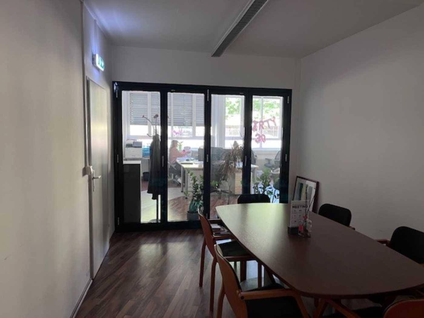 Besprechungsraum mit Holztisch und Glaswand, die den Blick in ein angrenzendes Büro freigibt.
