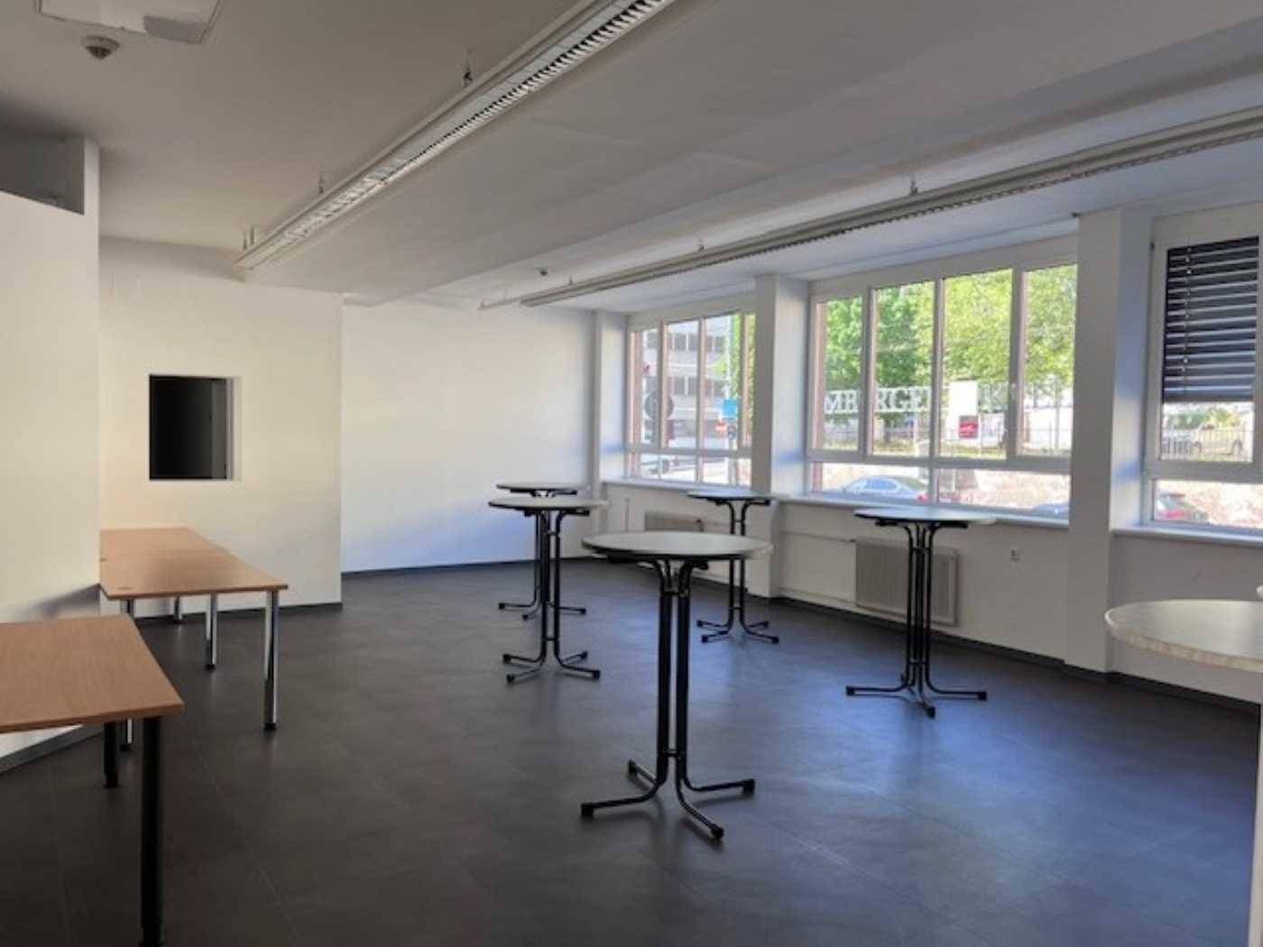 Großer Mehrzweckraum mit Stehtischen, Holztischen und großen Fenstern mit viel Tageslicht.