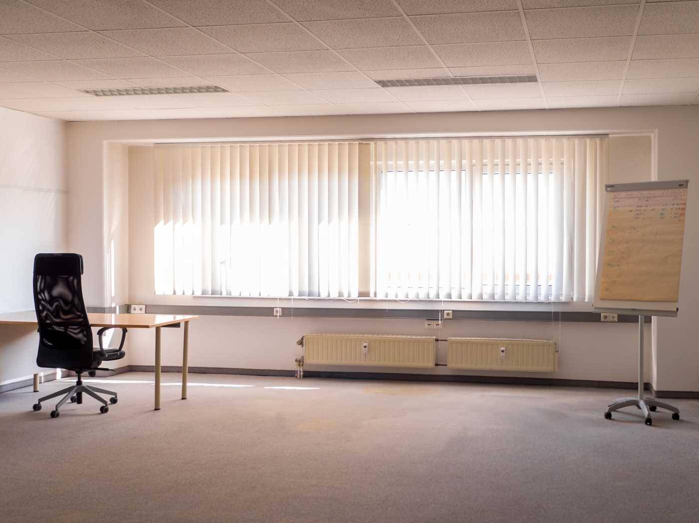 Leeres Büro mit Teppichboden, großen Fenstern mit Jalousien und einem Flipchart.