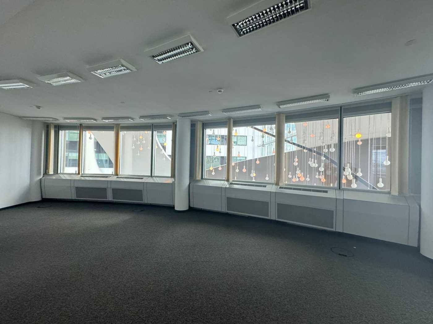 Leerer Büroraum mit Teppichboden und Fenstern, die einen Blick auf das Gebäudeinnere bieten.