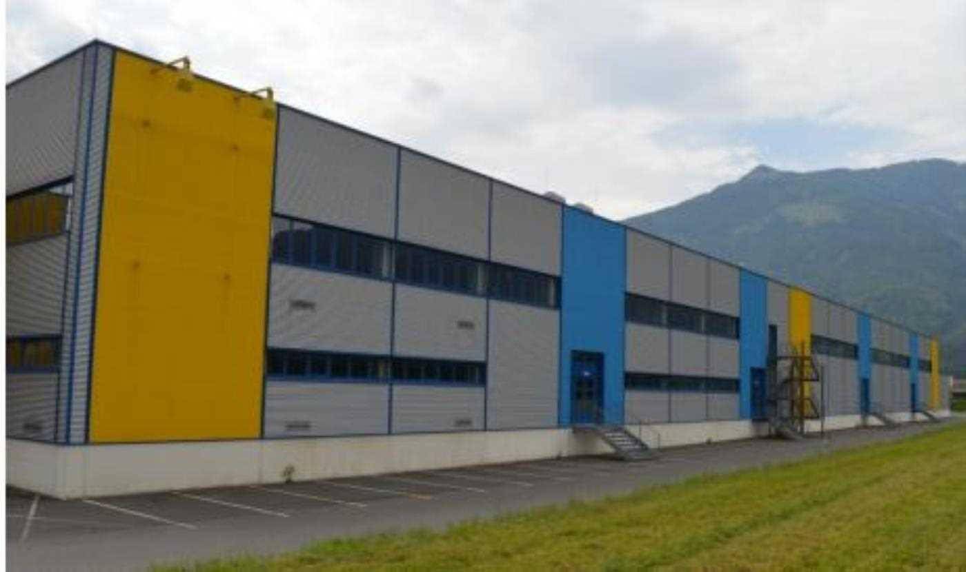 Modernes Industriegebäude mit grauer, blauer und gelber Fassade vor einer Bergkulisse.