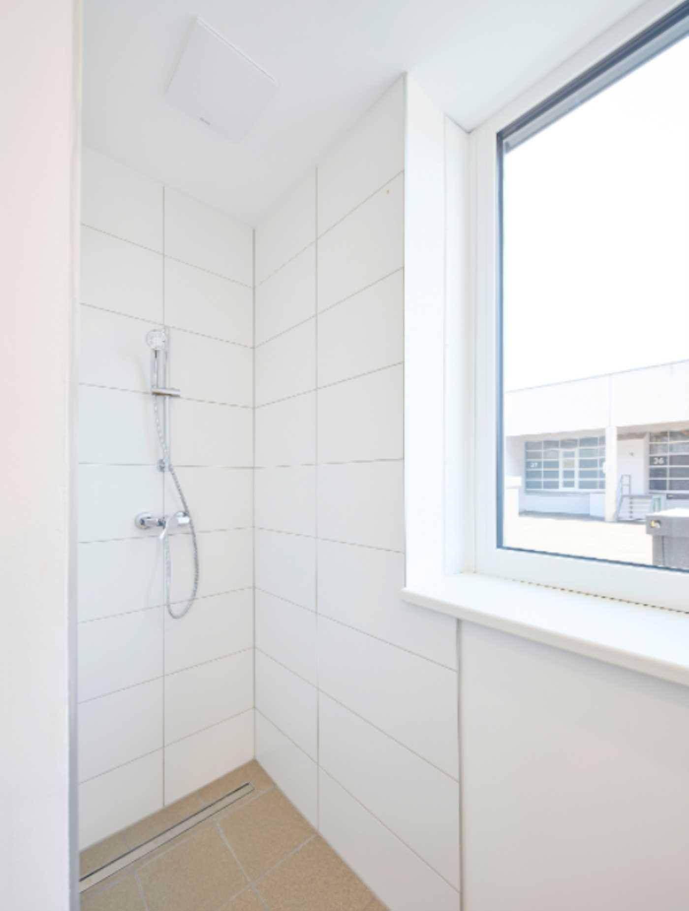 Einfache Duschkabine mit weißen Fliesen, Duschkopf und einem Fenster für Belüftung.