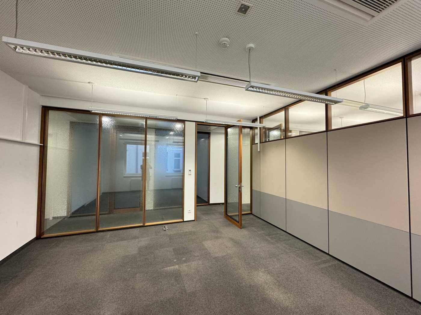 Geräumiges Büro mit Teppichboden, Glaswänden und abgehängter Decke mit integrierter Beleuchtung.
