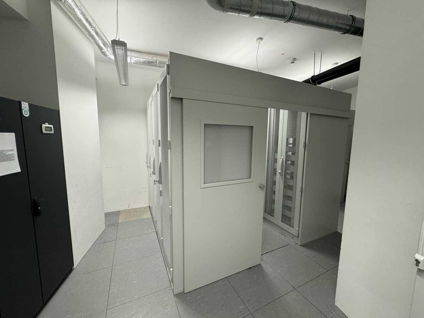 Technischer Raum mit Serverracks, Fliesenboden und sichtbaren Lüftungsrohren an der Decke.