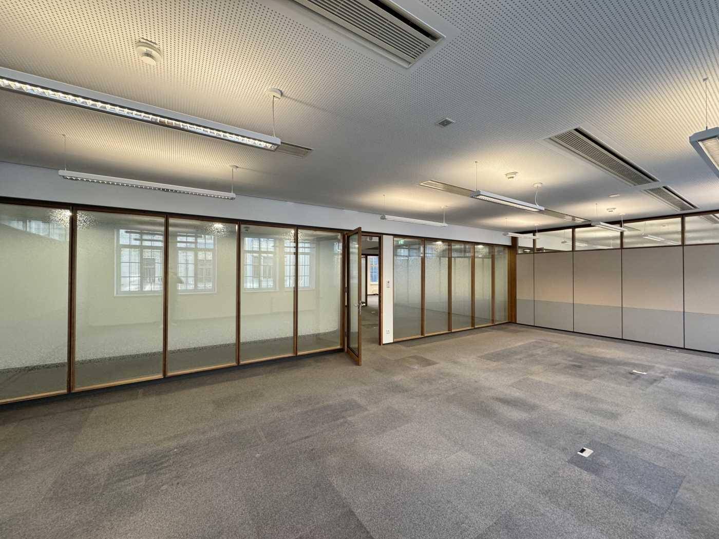Geräumiges Büro mit Teppichboden, Glaswänden und abgehängter Decke mit integrierter Beleuchtung.