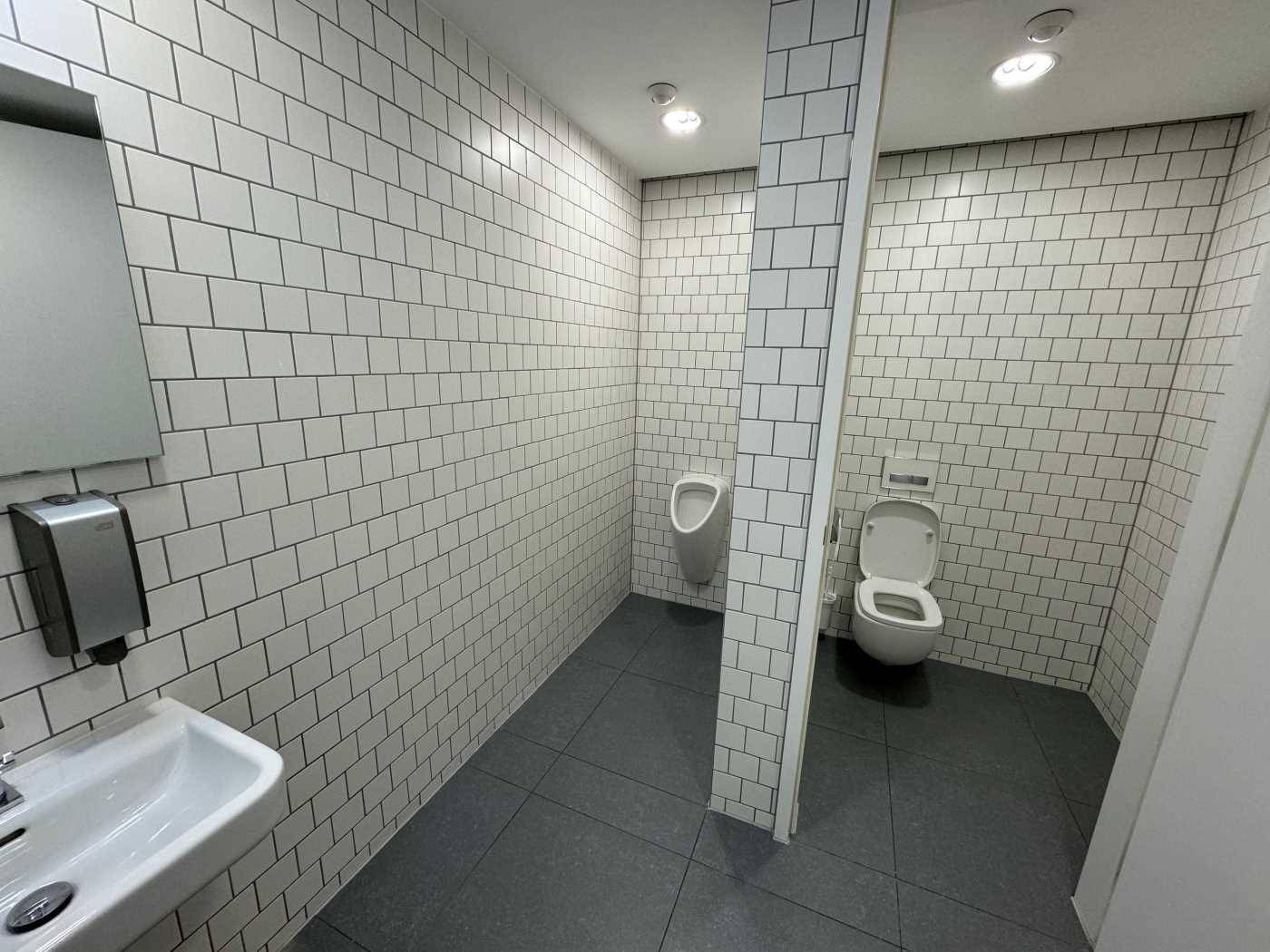 Modernes WC mit weißen Fliesen, Urinal und Waschbecken in sauberem Zustand.