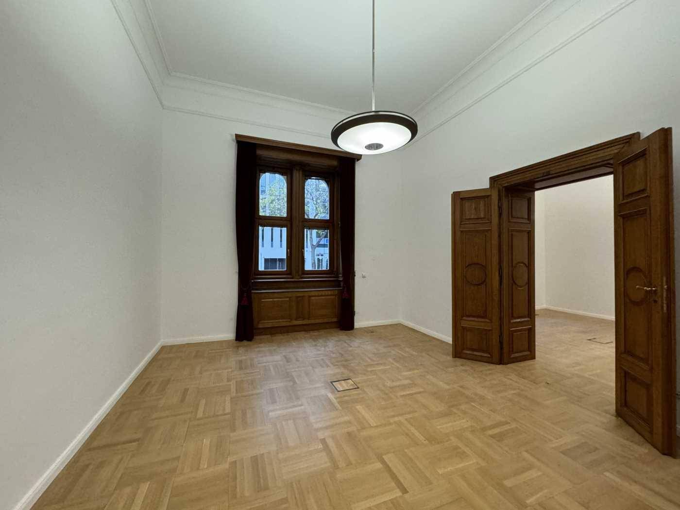 Heller Büroraum mit Parkettboden, hohen Decken und einem großen Fenster mit dunklen Vorhängen.