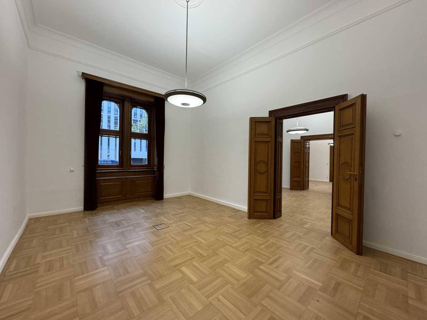 Geräumiger Büroraum mit Parkettboden, hohen Decken und einem großen Fenster sowie mehreren Holztüren.