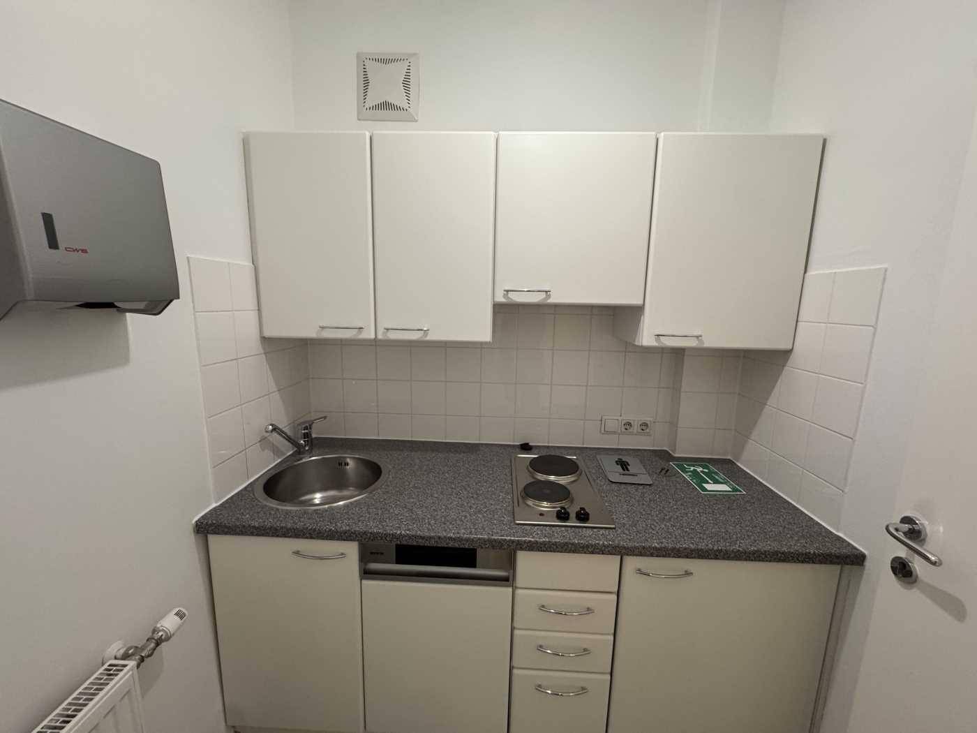 Kleine Einbauküche mit weißen Schränken, Spüle, Kochfeld und Dunstabzugshaube.