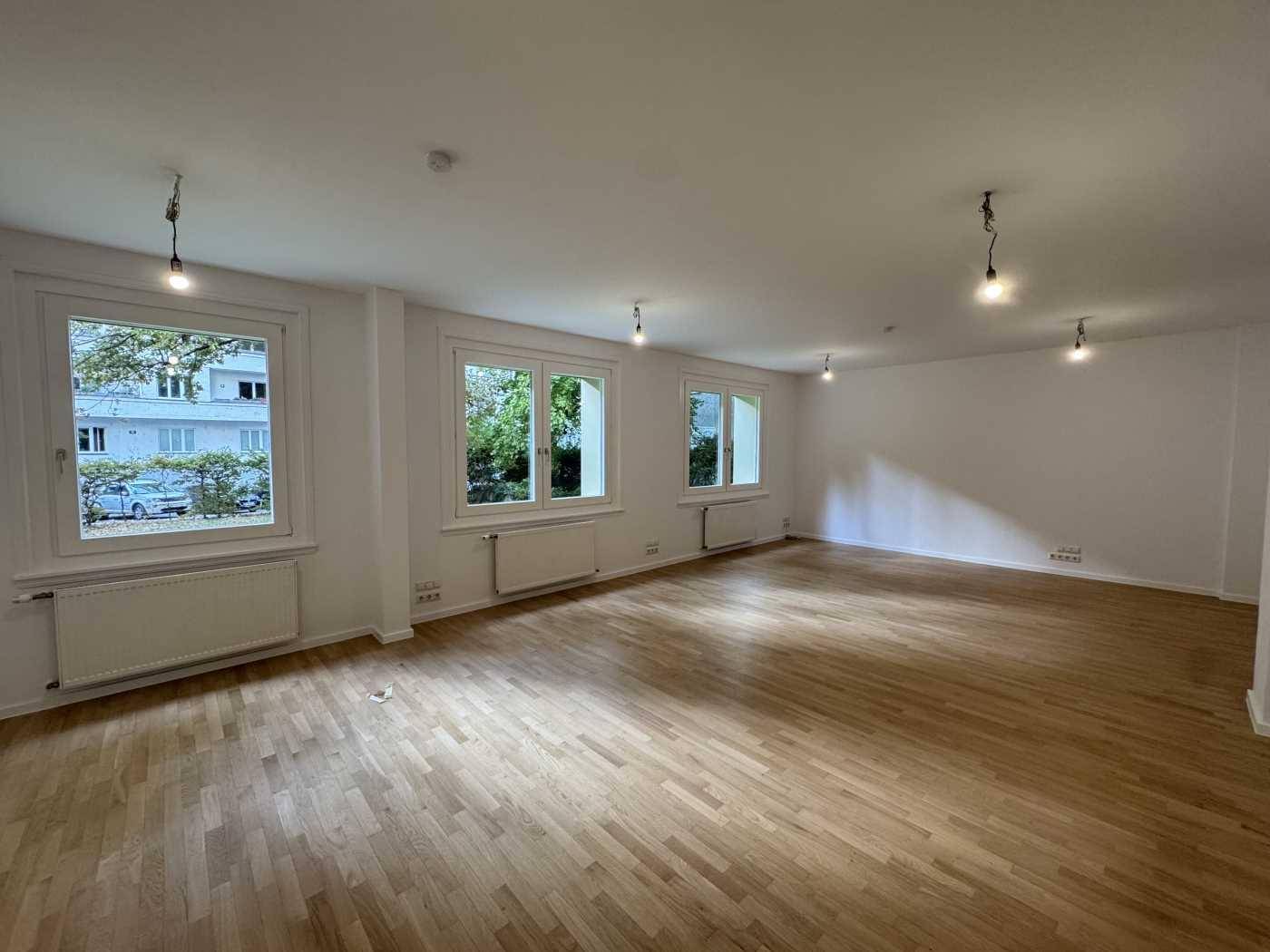 Geräumiger, leerer Büroraum mit Holzboden und mehreren Fenstern, die viel Tageslicht hereinlassen.