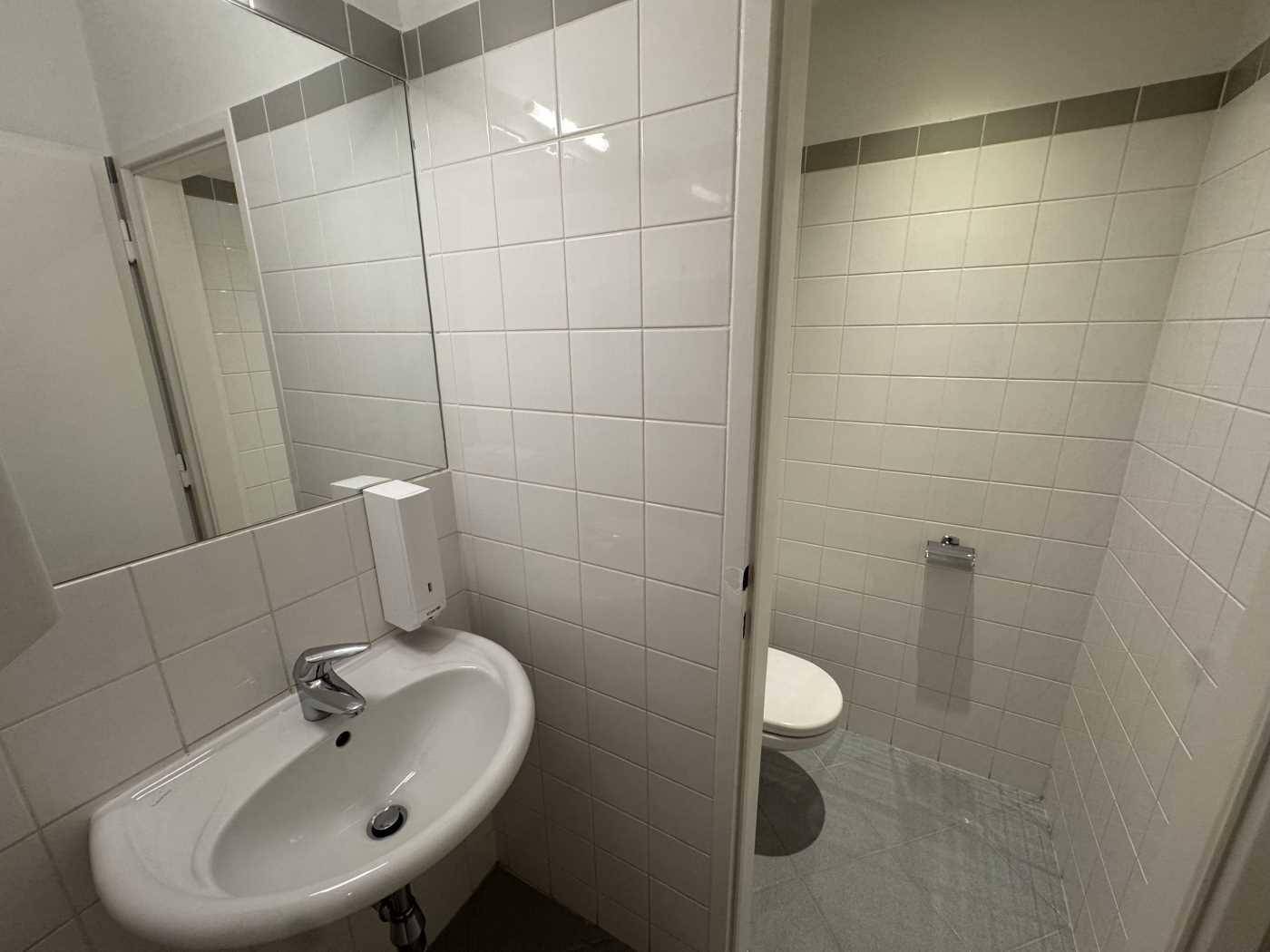 Kleiner Toilettenraum mit weiß gefliesten Wänden, Waschbecken und Spiegel.