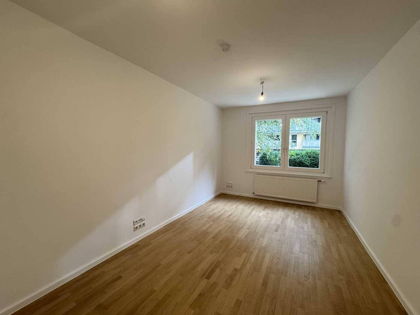 Leerer Büroraum mit Holzboden und einem Fenster, das den Blick ins Grüne bietet.